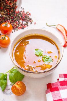 Delicious tomato soup with meat in a white bowl on a wooden table with fresh cherry tomatoes, basil leaves, cut chili pepper and red gingham kitchen towel. Ingredients for soup. Top view