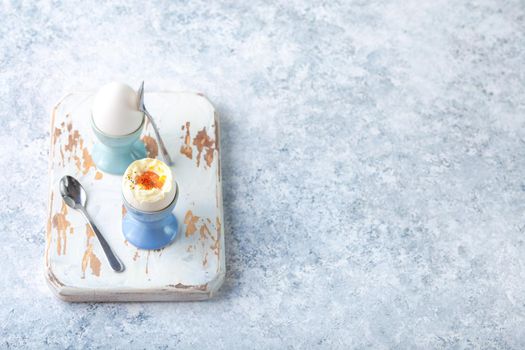 Fresh soft boiled eggs in stands, spoons, wooden cooking board, white concrete rustic background. Top view. Soft eggs, healthy breakfast. Space for text. Selective focus. Egg protein fitness breakfast