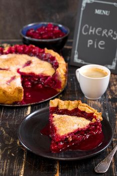 Slice of homemade cherry pie, cup of coffee, bowl with cherries and menu chalkboard on the black wooden table