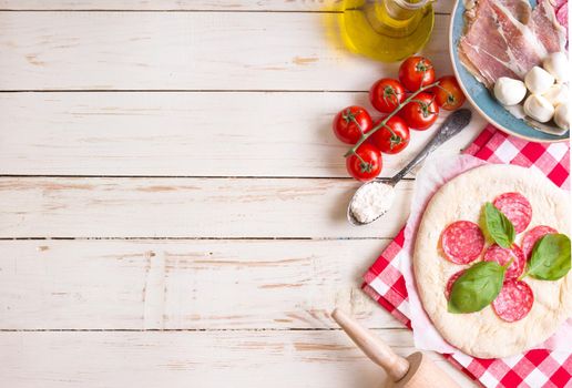 Pizza making background. Ingredients for making pizza. Space for text. Pizza dough, flour, cheese, mozzarella, tomatoes, basil, pepperoni, olives and rolling pin over white wooden background. Top view