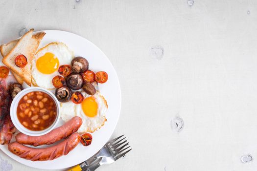 Full english breakfast with fried eggs, tomatoes, sausages, bacon, mushrooms, toasts and beans. Breakfast on a white plate with forks on the white wooden table. Space for text. Background