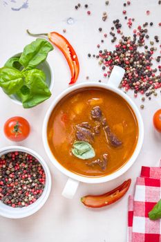 Delicious tomato soup with meat on a white rustic wooden table with fresh cherry tomatoes, basil leaves, cut chili pepper and red gingham kitchen towel. Ingredients for soup. Top view