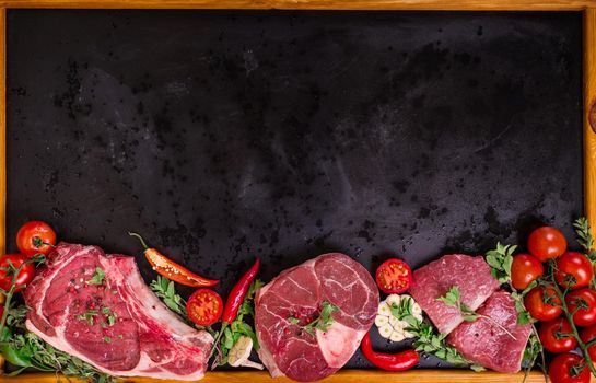 Raw juicy meat steaks on a black chalk board background with wooden frame. Rib eye steak on the bone, veal shank (ossobuco), fillet with cherry tomatoes, hot pepper and herbs. Space for text