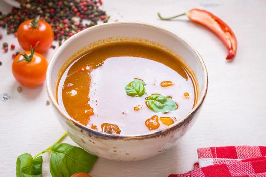 Delicious tomato soup with meat in a white bowl on a wooden table with fresh cherry tomatoes, basil leaves, cut chili pepper and red gingham kitchen towel. Ingredients for soup. Top view