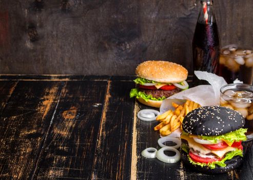 Set of black burgers with meat patty, cheese, tomatoes, mayonnaise, french fries and glass of cold cola soda with ice. Dark wooden rustic background. Space for text. Modern fast food lunch frame