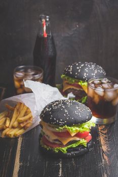 Set of black burgers with meat patty, cheese, tomatoes, mayonnaise, french fries in a paper cup and glass of cold cola soda with ice. Dark wooden rustic table. Modern fast food lunch. Toned image