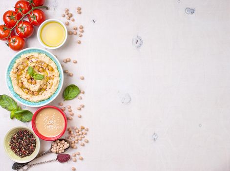 Bowl with hummus, chickpea, tahini, olive oil, sesame seeds, cherry tomatoes and herbs on white rustic wooden background. Space for text. Food frame. Middle eastern cuisine. Top view. Hummus background