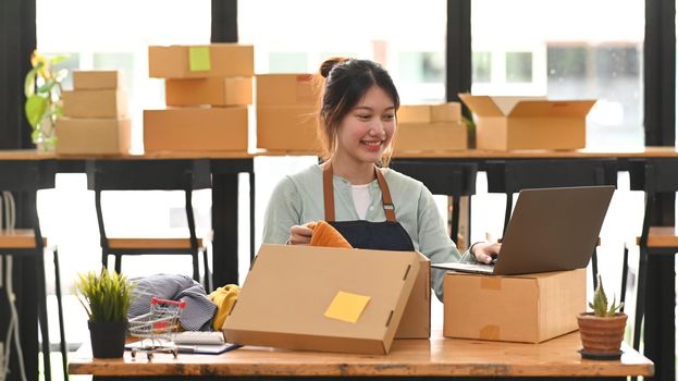 Smiling female small business owner using laptop during packing product in cardboard box for delivery to customers. Online selling, e-commerce.