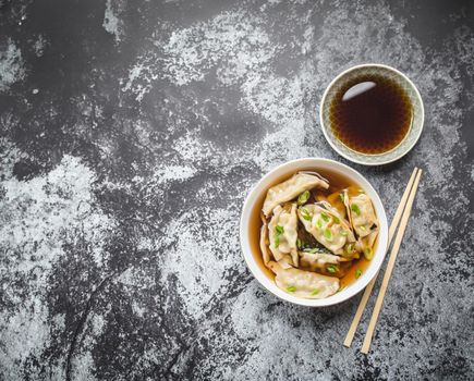 Asian dumplings in broth, bowl, chopsticks, soy sauce, rustic stone background. Top view. Space for text. Chinese dumplings for dinner. Closeup. Traditional Asian/Chinese cuisine. Overhead. Soup