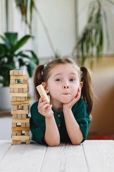 Portrait of beautiful cute little girl in green dress with ponytails on her head. showing a wooden block at a camera and wooden tower standing on a table.