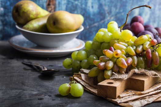 Fresh ripe grapes on wooden cutting board, pears and plums in bowls, wooden rustic background. Space for text. Close up. Organic natural fruits. Autumn/fall setting. Harvest/crop/garden concept