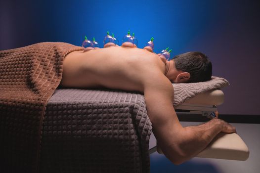 Close-up of a man's back with glass vacuum cans installed in a dark room of a spa salon.