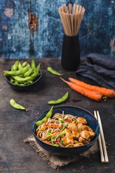 Asian noodles with chicken, vegetables in bowl, rustic wooden background. Soba noodles, teriyaki sauce chicken, edamame beans, sesame, chopsticks. Closeup. Asian style dinner. Chinese/Japanese noodles