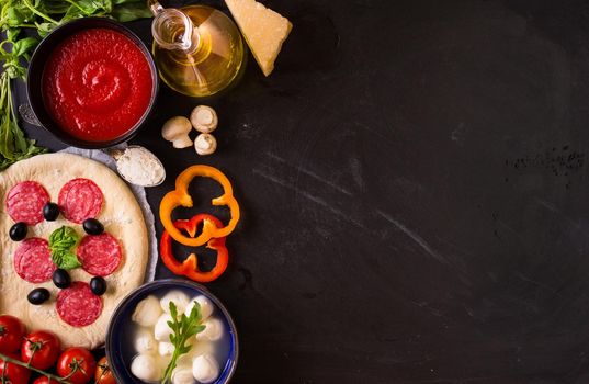 Pizza making background. Ingredients for making pizza. Space for text. Pizza dough, flour, cheese, mozzarella, tomatoes, basil, pepperoni, olives and rolling pin over black chalkboard. Top view