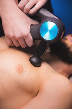 Medium shot of caucasian professional male massage therapist getting pectoral muscles with massage gun percussion tool of muscular athlete, in spa treatments, lying on back in massage table