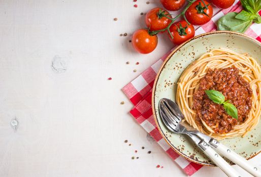 Italian pasta bolognese. Spaghetti with meat and tomato sauce in a plate with Italian tablecloth on a wooden white background. With fresh cherry tomatoes, basil. Food frame. Space for text. Top view