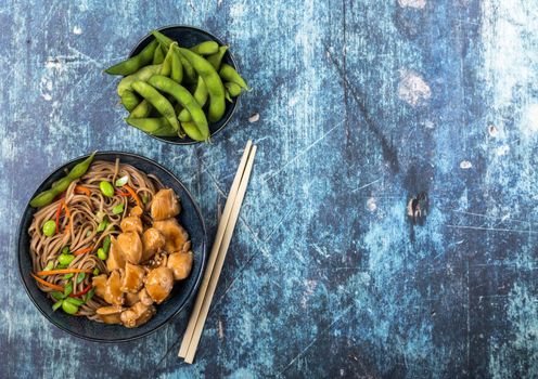 Asian noodles with chicken, vegetables, bowl, rustic wooden blue background. Space for text. Top view. Soba noodles, teriyaki chicken, edamame, chopsticks. Asian style dinner. Chinese/Japanese noodles