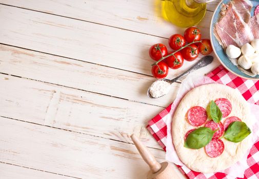 Pizza making background. Ingredients for making pizza. Space for text. Pizza dough, flour, cheese, mozzarella, tomatoes, basil, pepperoni, olives and rolling pin over white wooden background. Top view