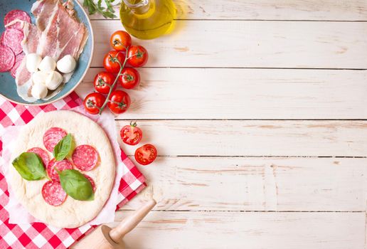 Pizza making background. Ingredients for making pizza. Space for text. Pizza dough, flour, cheese, mozzarella, tomatoes, basil, pepperoni, olives and rolling pin over white wooden background. Top view