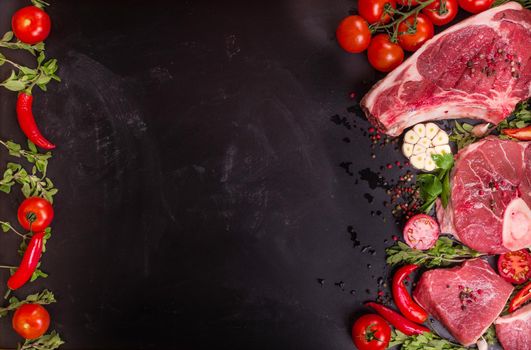 Raw juicy meat steaks ready for roasting on a black chalk board background. Rib eye steak on the bone, veal shank (ossobuco), fillet with cherry tomatoes, hot pepper and herbs. Space for text