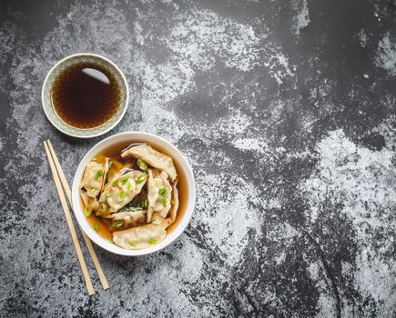 Asian dumplings in broth, bowl, chopsticks, soy sauce, rustic stone background. Top view. Space for text. Chinese dumplings for dinner. Closeup. Traditional Asian/Chinese cuisine. Overhead. Soup