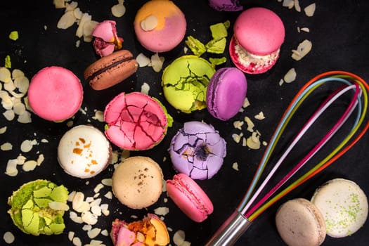 Assorted colorful french macarons and almond flakes with a whisk on a black background. Closeup. Top view. Concept of the baking macarons