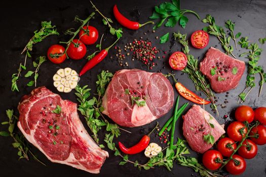 Different types of steaks set. Assorted raw meat on a black chalk board background. Rib eye steak on the bone, veal shank (ossobuco), fillet with cherry tomatoes, hot pepper and herbs.