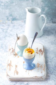Fresh soft boiled eggs in stands, spoons, white wooden cooking board, white concrete rustic background. Soft eggs for healthy breakfast. Jar with milk. Selective focus. Egg protein fitness breakfast