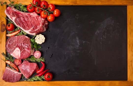 Raw juicy meat steaks on a black chalk board background with wooden frame. Rib eye steak on the bone, veal shank (ossobuco), fillet with cherry tomatoes, hot pepper and herbs. Space for text