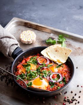 Shakshuka with pita bread in a pan. Middle eastern traditional dish. Fried eggs with tomatoes, bell pepper, vegetables and herbs. Shakshouka on a table. Sunny side up eggs. Homemade. Selective focus