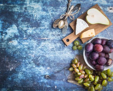 Fresh ripe pear, assorted cheeses platter, grapes, plums in bowl, blue wooden rustic background. Space for text. Top view. Fruits, Camembert, Emmental cheeses. Delicacy food. Snack/appetizer