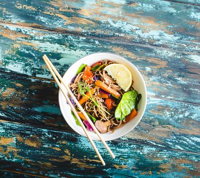 Asian noodles with vegetables, bowl, rustic wooden old background. From above. Top view. Soba noodles, vegetables, mushrooms, chopsticks. Vegetarian/Vegan noodles. Asian style dinner with noodles