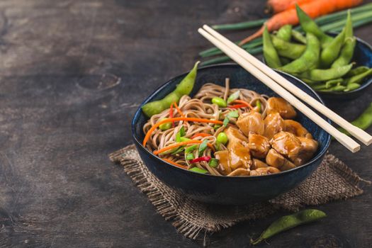 Asian noodles with chicken, vegetables in bowl, rustic wooden background. Space for text. Soba noodles, teriyaki chicken, edamame, chopsticks. Closeup. Asian style dinner. Chinese/Japanese noodles
