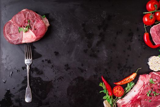 Raw juicy meat steaks ready for roasting on a black chalk board background. Meat on a fork. Veal shank (ossobuco), rib eye steak on the bone, fillet with cherry tomatoes, hot pepper and herbs.