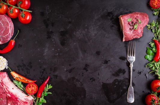 Raw juicy meat steaks ready for roasting on a black chalk board background. Meat on a fork. Veal shank (ossobuco), rib eye steak on the bone, fillet with cherry tomatoes, hot pepper and herbs.