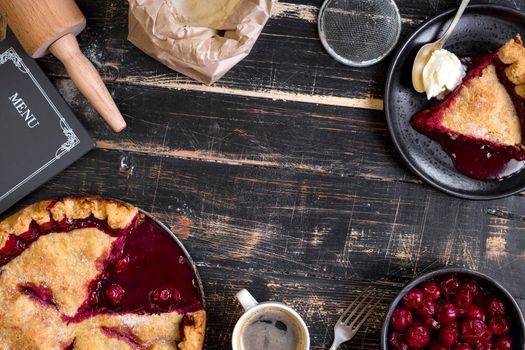 Homemade sliced cherry pie, bag with flour, rolling pin on the black wooden table. Baking background. Top view