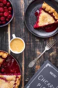Homemade sliced cherry pie with flaky crust, cup of coffee, bowl with cherries and menu chalkboard on the black wooden table. Top view