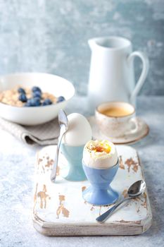 Fresh soft boiled eggs, oatmeal with blueberries, coffee cup, milk jug. White concrete rustic background. Soft eggs, healthy breakfast. Selective focus. Tasty light fitness breakfast concept. Closeup