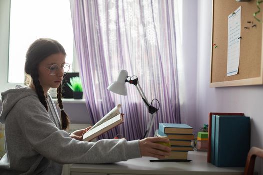 A teenage girl is sitting at a desk in her room. Girl reaches for an apple while reading her school reading. Young girl wearing glasses with braids.