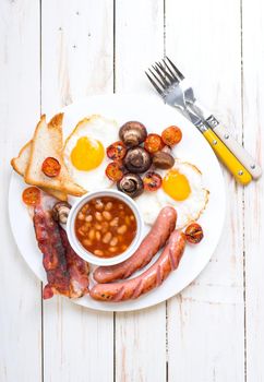 Full english breakfast with fried eggs, tomatoes, sausages, bacon, mushrooms, toasts and beans. Breakfast on a white plate with forks on the white wooden table. Top view