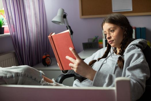 A schoolgirl is reading a highly engaging adventure book. The brunette has her glasses on and her hair styled in two braids. Book lover.