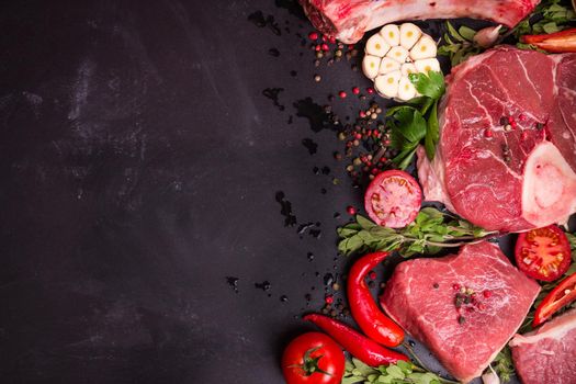 Raw juicy meat steaks ready for roasting on a black chalk board background. Rib eye steak on the bone, veal shank (ossobuco), fillet with cherry tomatoes, hot pepper and herbs. Space for text