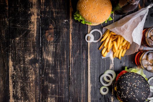 Set of black burgers with meat patty, cheese, tomatoes, mayonnaise, french fries and glass of cold cola soda with ice from above. Dark wooden rustic background. Space for text. Top view