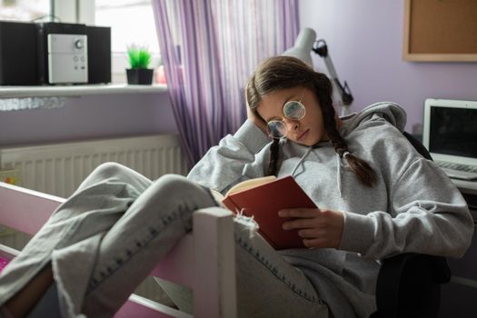 A young girl completely absorbed in reading a book. The teenager has her glasses on and her hair styled in two braids.