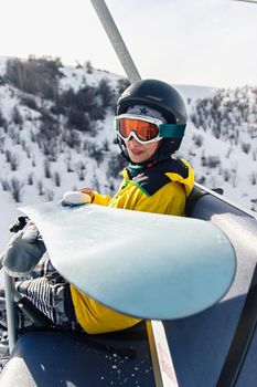 Young woman snowboarder sitting on ski lift holding her snowboard, she's looking at camera