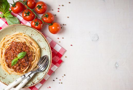 Italian pasta bolognese. Spaghetti with meat and tomato sauce in a plate with Italian tablecloth on a wooden white background. With fresh cherry tomatoes, basil. Food frame. Space for text. Top view