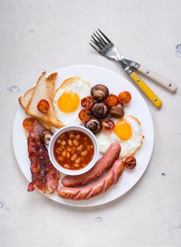 Full english breakfast with fried eggs, tomatoes, sausages, bacon, mushrooms, toasts and beans. Breakfast on a white plate with forks on the white wooden table. Top view