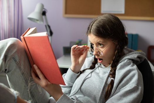 A student is surprised by a twist she read in a book. The brunette has her glasses on and her hair styled in two braids.