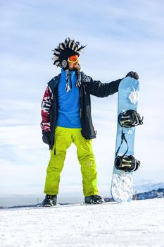 Bearded happy snowboarder in a ski mask with goggles and a fur big mohawk hat on a background of a sky and winter snowy mountains