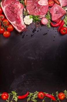 Raw juicy meat steaks ready for roasting on a black chalk board background. Rib eye steak on the bone, veal shank (ossobuco), fillet with cherry tomatoes, hot pepper and herbs. Space for text